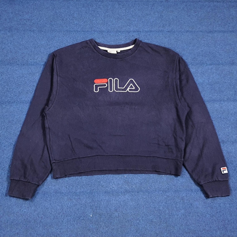 Puede incluir: Sudadera azul marino con el nombre de la marca "FILA" en letras rojas y blancas en el pecho. La sudadera tiene cuello redondo, mangas largas y un peque&ntilde;o logotipo en el pu&ntilde;o de la manga izquierda. La prenda es de material suave.