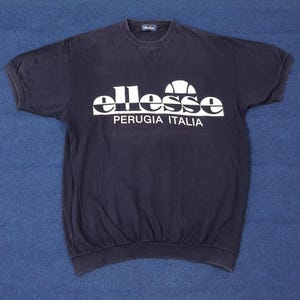 May include: A navy blue short-sleeved sweatshirt featuring the white brand name "ellesse" and the text "PERUGIA ITALIA". The sweatshirt has a crew neck and a relaxed fit, suitable for casual wear.