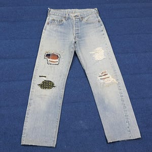 Könnte beinhalten: Hellblaue Jeans aus Denim mit Used-Details, einschließlich Rissen und Patches. Die Patches weisen Karomuster und geometrische Muster auf, die auf den Denim genäht sind. Die Jeans haben einen Knopfverschluss und ein klassisches Fünf-Taschen-Design.