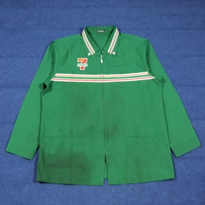 Puede incluir: Chaqueta de trabajo verde de manga larga con cremallera completa. La chaqueta presenta una franja blanca y roja en el pecho y el logotipo de 7-Eleven en el pecho izquierdo. El cuello tiene rayas blancas y verdes.