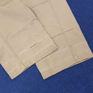 May include: Close-up of tan-colored pants with visible stitching and a distressed, ripped detail near the hem. The fabric appears to be a sturdy cotton or blend, with a slightly worn look. The pants are folded, showing the leg seams.