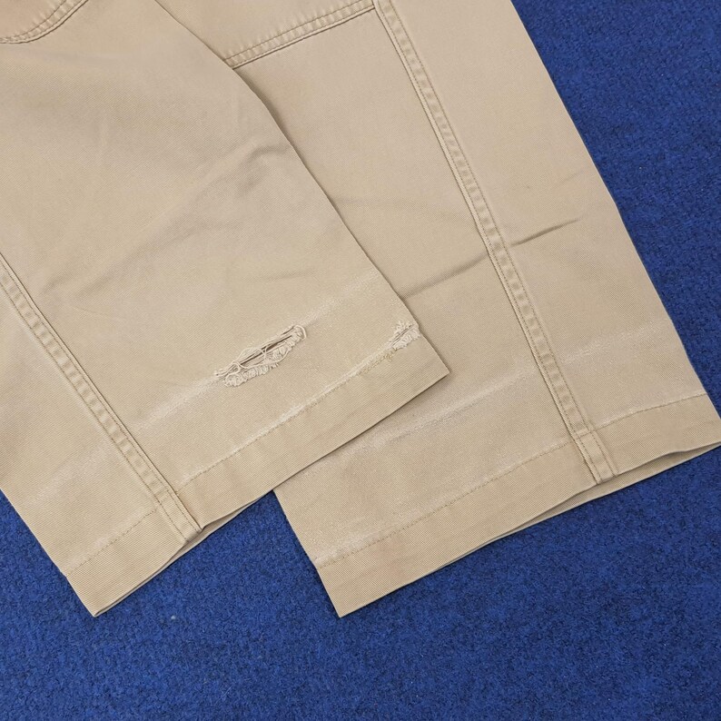 May include: Close-up of tan-colored pants with visible stitching and a distressed, ripped detail near the hem. The fabric appears to be a sturdy cotton or blend, with a slightly worn look. The pants are folded, showing the leg seams.