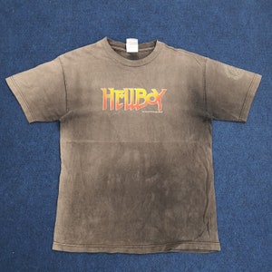 May include: A brown t-shirt with a faded graphic of the word "HELLBOY" in yellow and orange letters. The shirt has a distressed look and is made of a soft, comfortable material.
