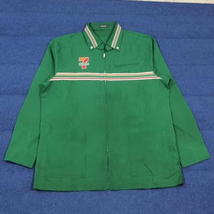 Puede incluir: Chaqueta verde con cremallera y el logotipo "7-Eleven" en el pecho. La chaqueta tiene una franja blanca, naranja y roja en el pecho y en el cuello. Dos bolsillos grandes están en la parte inferior delantera. La chaqueta está hecha de un material duradero.