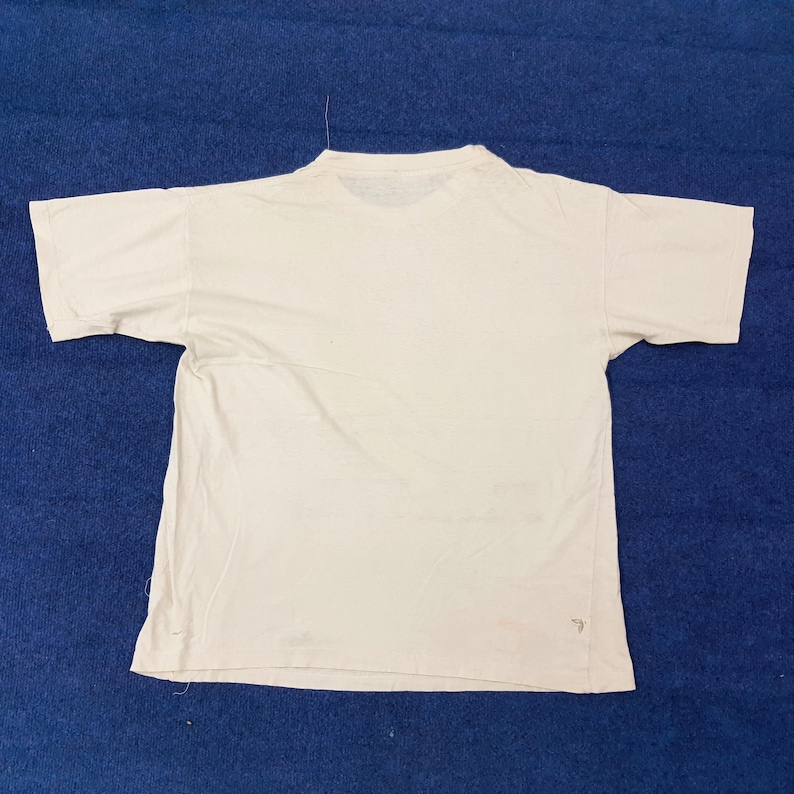 May include: A plain white t-shirt with short sleeves, laid flat on a blue surface. The shirt has a crew neck and appears to be made of a soft, lightweight fabric. The shirt is slightly wrinkled, suggesting it has been worn.