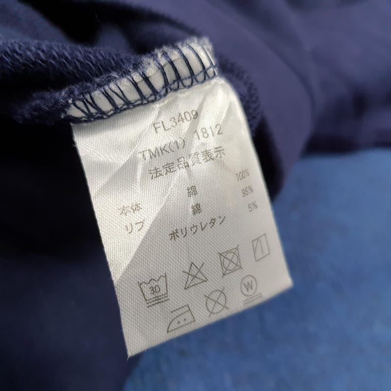 Puede incluir: Primer plano de una etiqueta de prenda azul marino con texto y s&iacute;mbolos. La etiqueta incluye los n&uacute;meros "FL3409" y "TMK(1) 1812". La etiqueta tambi&eacute;n enumera los materiales: algod&oacute;n y poliuretano.
