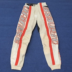 Puede incluir: Pantalones de moto blanquecinos con rayas rojas y la palabra "DAINESE" impresa verticalmente en cada pierna. Los pantalones tienen zonas acolchadas en las rodillas y una cintura negra. Los pu&ntilde;os son blancos.