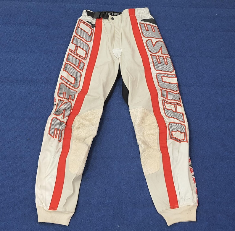 Puede incluir: Pantalones de moto blanquecinos con rayas rojas y la palabra "DAINESE" impresa verticalmente en cada pierna. Los pantalones tienen zonas acolchadas en las rodillas y una cintura negra. Los pu&ntilde;os son blancos.