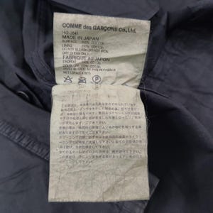 May include: Close-up of a garment tag from COMME des GAR&Ccedil;ONS. The tag is white with black text, listing the garment's composition as 100% cotton and the manufacturing location as Japan. The tag also includes care instructions and Japanese text.