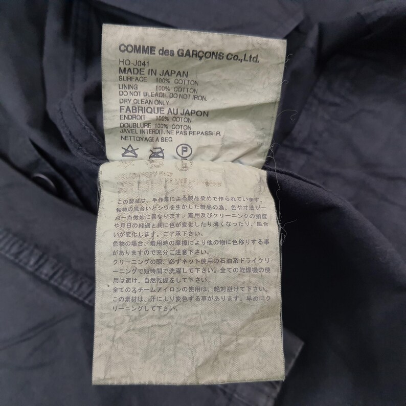 May include: Close-up of a garment tag from COMME des GAR&Ccedil;ONS. The tag is white with black text, listing the garment's composition as 100% cotton and the manufacturing location as Japan. The tag also includes care instructions and Japanese text.