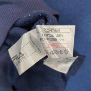 Puede incluir: Primer plano de una prenda azul marino con una etiqueta blanca. La etiqueta dice "CONTAIN" con los porcentajes de algod&oacute;n y poli&eacute;ster. La marca "FILA QUALITY" tambi&eacute;n es visible.