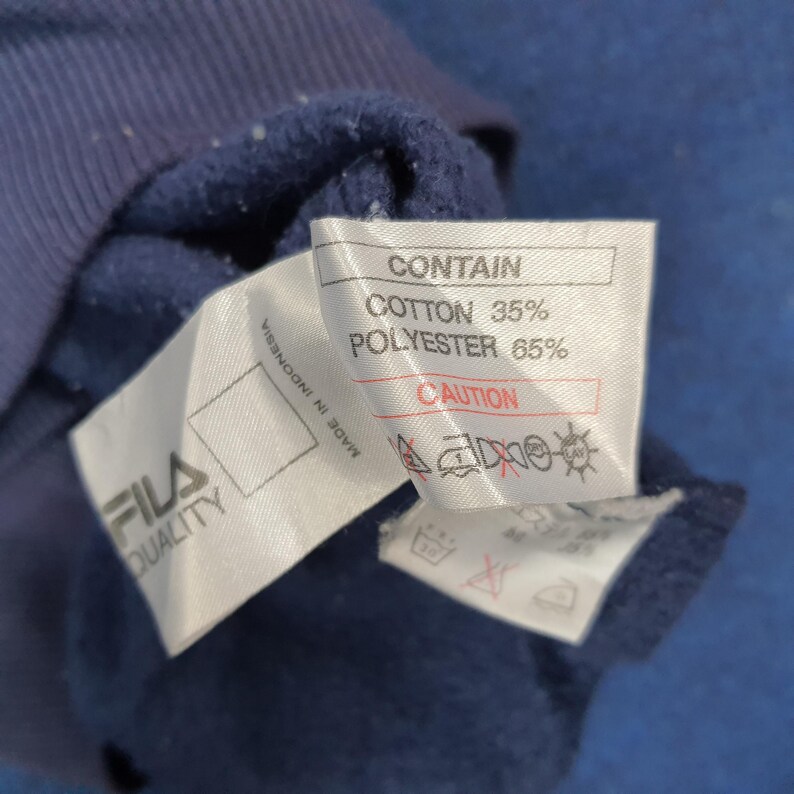 Puede incluir: Primer plano de una prenda azul marino con una etiqueta blanca. La etiqueta dice "CONTAIN" con los porcentajes de algod&oacute;n y poli&eacute;ster. La marca "FILA QUALITY" tambi&eacute;n es visible.