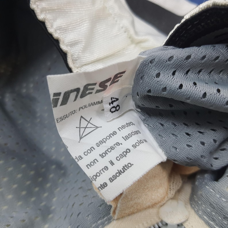 Puede incluir: Primer plano de una etiqueta blanca con la marca "Dainese" impresa en negro. La etiqueta tambi&eacute;n incluye instrucciones de lavado y la talla "48". La etiqueta est&aacute; adherida a una tela gris perforada.