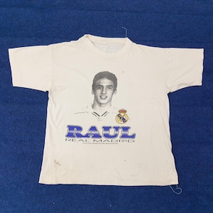 May include: White t-shirt featuring a portrait of Ra&uacute;l, a soccer player, with the text "RAUL REAL MADRID" in blue. The shirt has a crew neck and short sleeves. The Real Madrid crest is also visible.