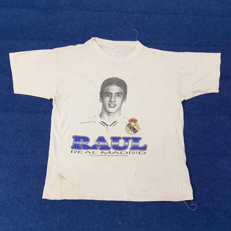 May include: White t-shirt featuring a portrait of Ra&uacute;l, a soccer player, with the text "RAUL REAL MADRID" in blue. The shirt has a crew neck and short sleeves. The Real Madrid crest is also visible.