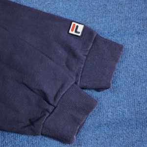 Puede incluir: Primer plano de una manga de sudadera azul marino. La manga presenta un pu&ntilde;o acanalado y un peque&ntilde;o parche rectangular con las letras "IL" en blanco sobre fondo rojo. La tela parece suave y ligeramente desgastada.