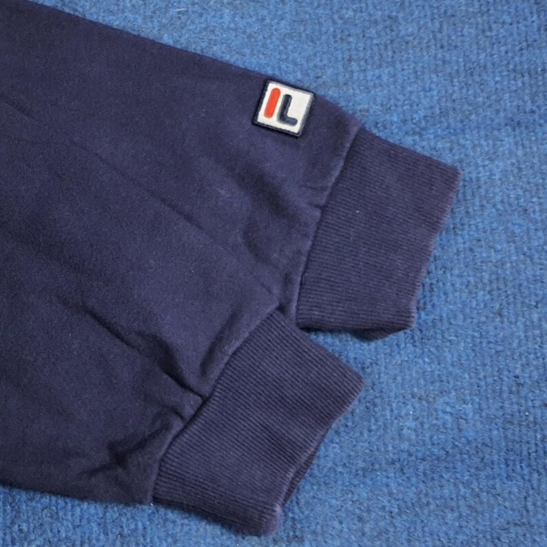 Puede incluir: Primer plano de una manga de sudadera azul marino. La manga presenta un pu&ntilde;o acanalado y un peque&ntilde;o parche rectangular con las letras "IL" en blanco sobre fondo rojo. La tela parece suave y ligeramente desgastada.