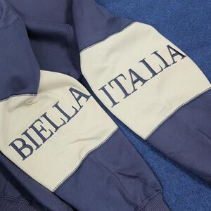 Puede incluir: Primer plano de una sudadera azul marino con paneles color crema en las mangas. Las mangas presentan el texto bordado "BIELLA ITALIA" en azul marino. La sudadera est&aacute; hecha de un material suave.
