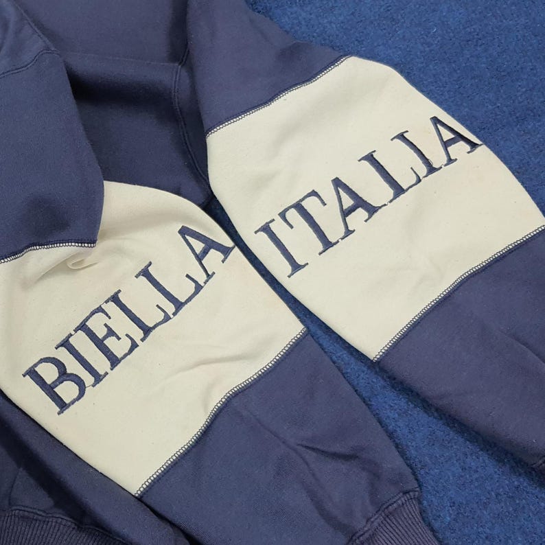 Puede incluir: Primer plano de una sudadera azul marino con paneles color crema en las mangas. Las mangas presentan el texto bordado "BIELLA ITALIA" en azul marino. La sudadera est&aacute; hecha de un material suave.