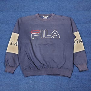 Puede incluir: Sudadera azul marino con la palabra "FILA" en blanco, contorneada en negro y una l&iacute;nea roja sobre la "A". Las mangas tienen un panel gris y blanco con las letras "ITA" y "LLA".