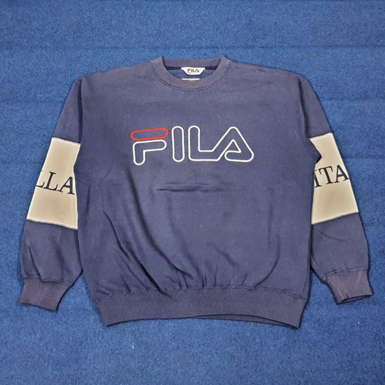 Puede incluir: Sudadera azul marino con la palabra "FILA" en blanco, contorneada en negro y una l&iacute;nea roja sobre la "A". Las mangas tienen un panel gris y blanco con las letras "ITA" y "LLA".