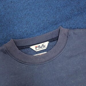 Puede incluir: Primer plano de una sudadera FILA azul marino. El cuello y los pu&ntilde;os son acanalados. El logotipo de FILA es visible en una etiqueta blanca. El fondo es una tela azul oscuro texturizada.