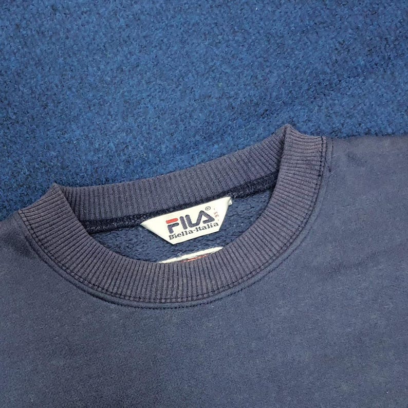 Puede incluir: Primer plano de una sudadera FILA azul marino. El cuello y los pu&ntilde;os son acanalados. El logotipo de FILA es visible en una etiqueta blanca. El fondo es una tela azul oscuro texturizada.