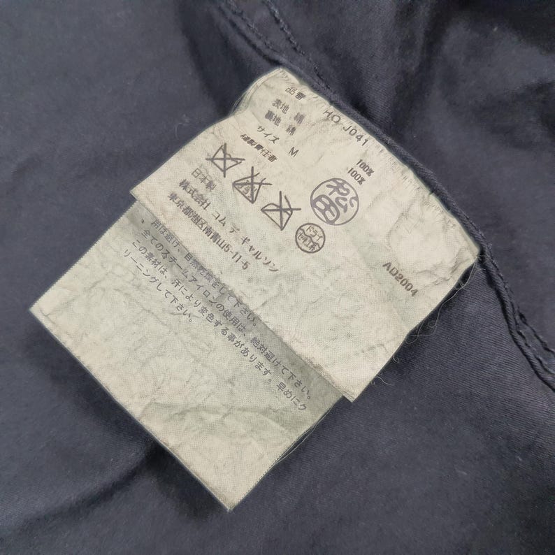 May include: Close-up of a navy blue garment with a tag attached. The tag has Japanese text, care symbols, and the text "AD2004". The tag is attached to the garment with a seam.