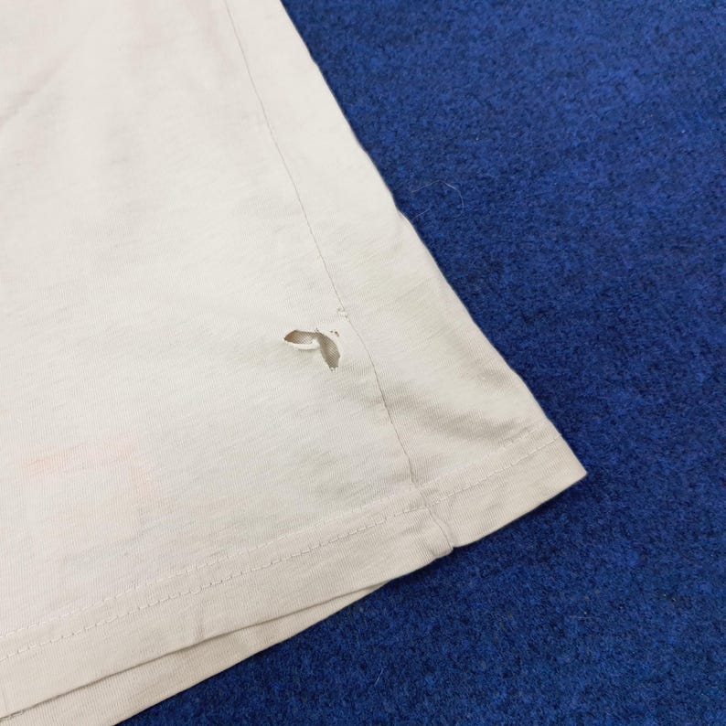 May include: A light beige t-shirt with a tear near the hem. The fabric appears to be a soft, lightweight material. The shirt is on a blue surface.