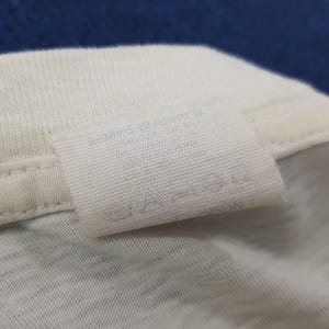 May include: Close-up of a white fabric tag sewn onto a white garment. The tag has faded text and symbols, likely care instructions. The fabric appears to be a soft, knit material. The background is a blurred blue.