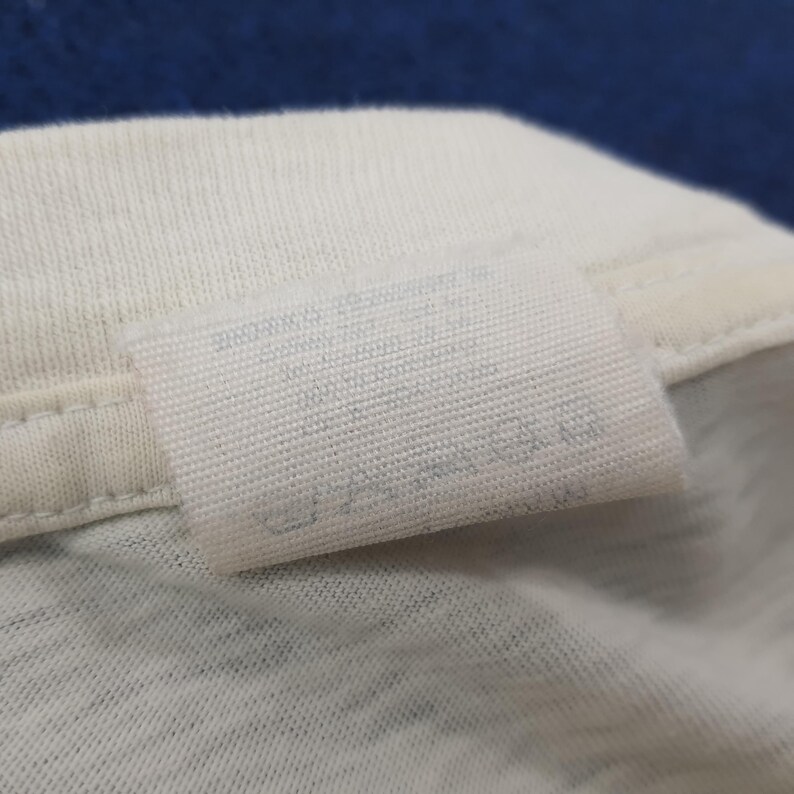 May include: Close-up of a white fabric tag sewn onto a white garment. The tag has faded text and symbols, likely care instructions. The fabric appears to be a soft, knit material. The background is a blurred blue.