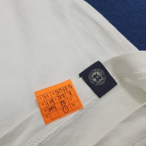 Puede incluir: Una camiseta blanca con una etiqueta azul que dice "Limited at the Estate of the King". La etiqueta tiene un peque&ntilde;o gr&aacute;fico de un p&aacute;jaro con una corona. Una etiqueta amarilla con una cuadr&iacute;cula de n&uacute;meros y letras est&aacute; unida a la camiseta.