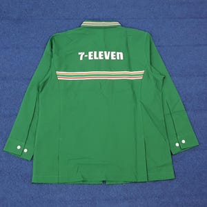 Puede incluir: Una camisa de trabajo verde de manga larga con el texto "7-ELEVEN" en blanco en la espalda. La camisa tiene una franja horizontal en rojo, blanco y verde. El cuello tiene un diseño a rayas verdes y blancas. Las mangas tienen botones blancos.