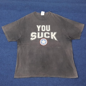 Puede incluir: Camiseta gris oscuro con las palabras "YOU SUCK" en letras blancas. Un emblema de estrella roja, blanca y azul está centrado debajo del texto. La camiseta tiene cuello redondo y mangas cortas.