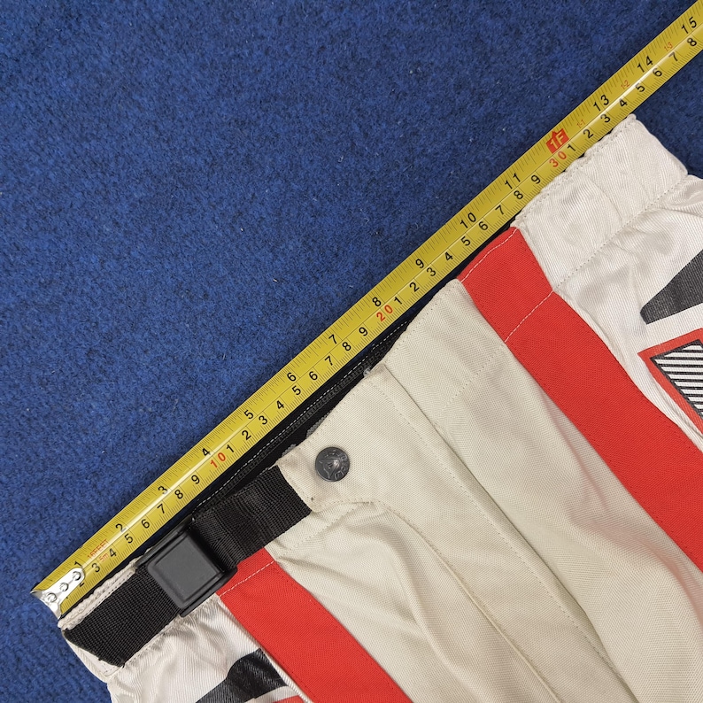 Puede incluir: Primer plano de pantalones de moto en crema, rojo y negro. Los pantalones tienen un cintur&oacute;n negro con hebilla y rayas rojas. Una cinta m&eacute;trica amarilla muestra las dimensiones en cent&iacute;metros.