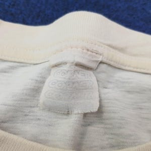 May include: Close-up of a cream-colored t-shirt collar and tag. The tag is rectangular with faded text. The shirt appears to be made of a soft, textured fabric. The collar is a lighter shade of cream.