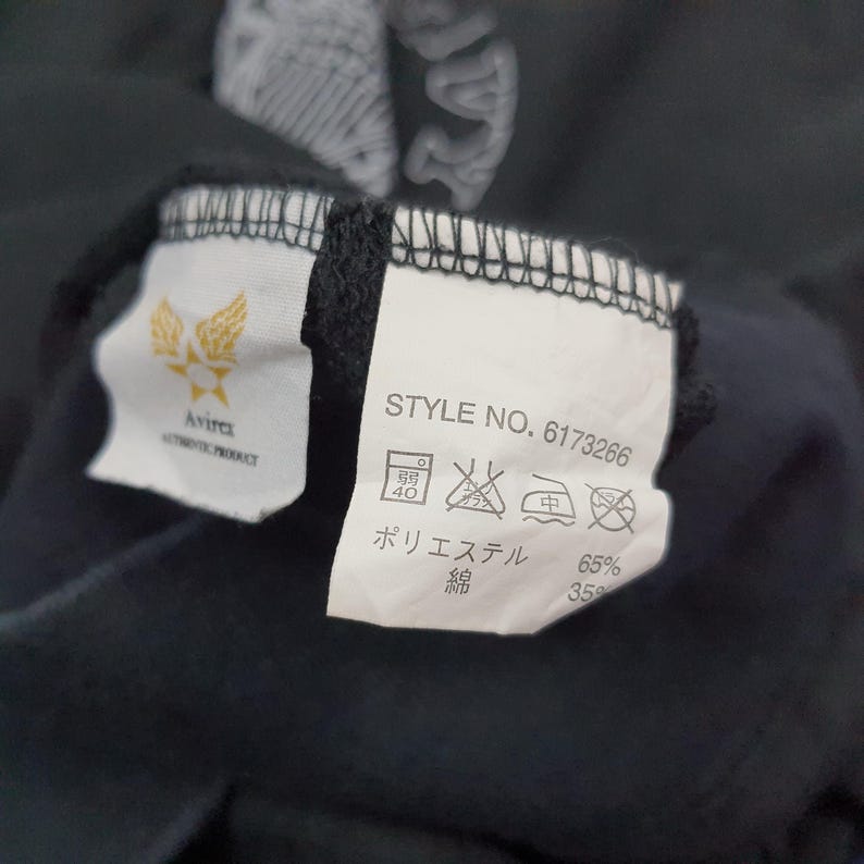 K&ouml;nnte beinhalten: Nahaufnahme eines schwarzen Kleidungsst&uuml;cks mit zwei angebrachten Etiketten. Eines zeigt ein goldenes Emblem und den Markennamen "Avirex". Das andere Etikett zeigt "STYLE NO. 6173266" und Pflegehinweise. Der Stoff besteht aus 65% Polyester und 35% Baumwolle.