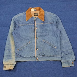 Puede incluir: Chaqueta vaquera vintage con cuello de pana marrón y cierre de botones. La chaqueta tiene dos bolsillos delanteros y el logo "Wrangler" es visible en el cuello. La chaqueta es de color azul medio.