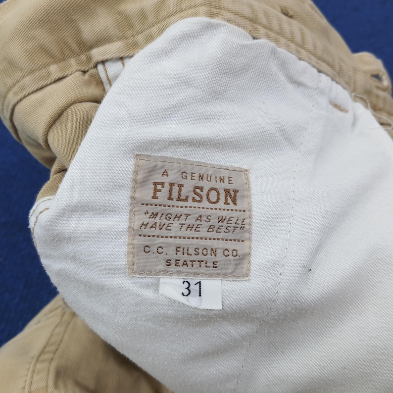 May include: Close-up of tan-colored pants with a white interior lining. A rectangular label reads "A GENUINE FILSON" with the text "MIGHT AS WELL HAVE THE BEST" and "C.C. FILSON CO. SEATTLE". A tag displays the number "31".