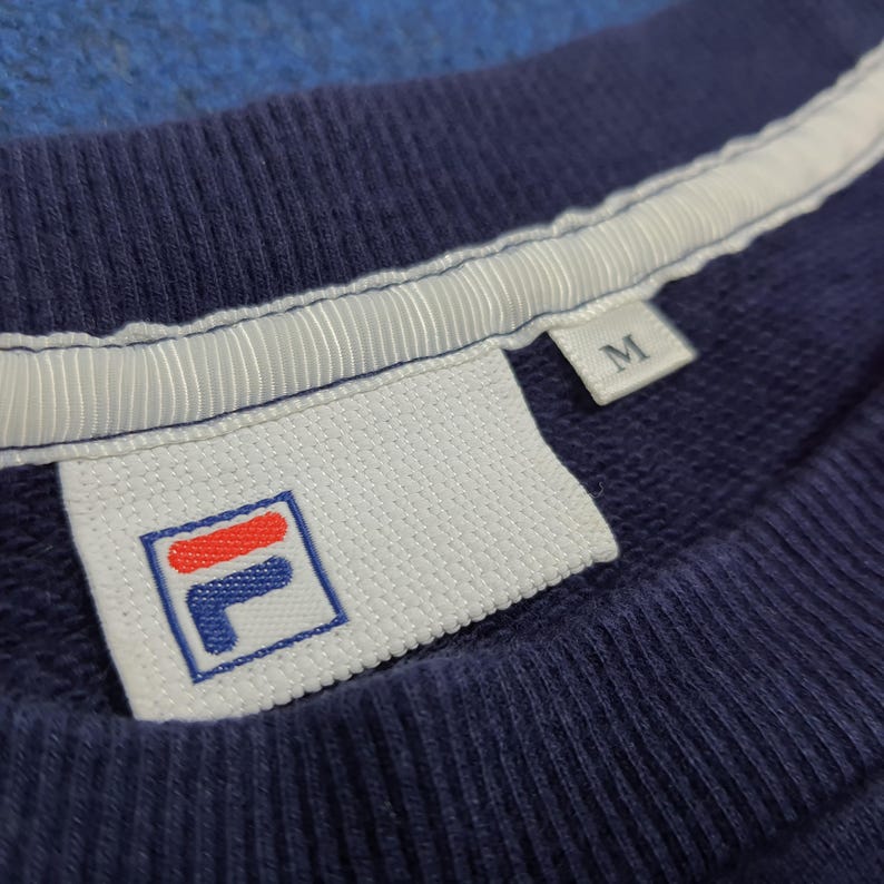 Puede incluir: Primer plano de una sudadera azul marino con una etiqueta blanca con el logotipo de la marca. El logotipo es un cuadrado con una 'L' azul y una franja roja. La etiqueta tambi&eacute;n tiene la letra 'M'. El cuello de la sudadera tiene una franja blanca.