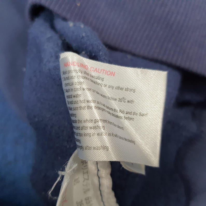 Puede incluir: Primer plano de una tela azul marino con una etiqueta blanca adjunta. La etiqueta tiene texto rojo y negro, incluyendo las palabras "HANDLING CAUTION" e instrucciones de cuidado. La tela parece ser parte de una prenda.