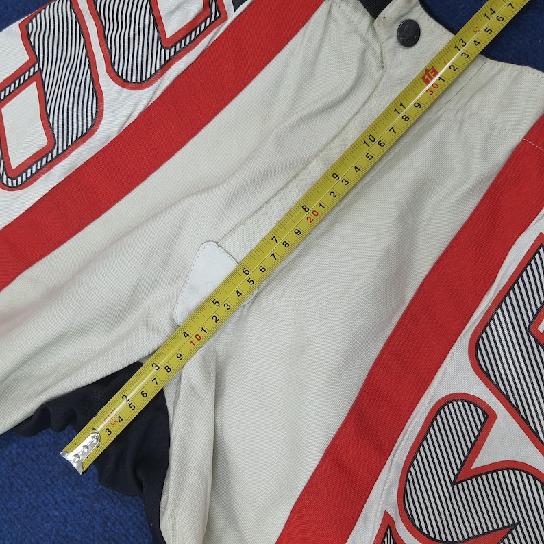 Puede incluir: Chaqueta color crema con detalles a rayas rojas y negras. Una cinta m&eacute;trica amarilla muestra medidas en cent&iacute;metros. La chaqueta tiene un cuello negro y un parche blanco. El dise&ntilde;o incluye elementos gr&aacute;ficos audaces.