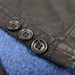 May include: Close-up of three black buttons sewn onto a dark fabric. The buttons are round with four holes each. The fabric appears to be a dark, possibly black, material with visible stitching. The background is a textured blue fabric.