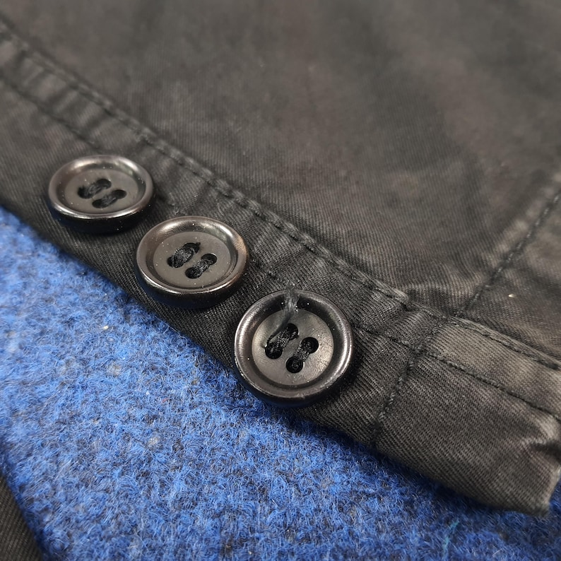 May include: Close-up of three black buttons sewn onto a dark fabric. The buttons are round with four holes each. The fabric appears to be a dark, possibly black, material with visible stitching. The background is a textured blue fabric.