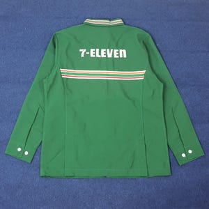 Puede incluir: Chaqueta verde de manga larga con el texto "7-ELEVEN" en blanco en la espalda. La chaqueta tiene un diseño de rayas blancas, rojas y naranjas en la espalda. La chaqueta tiene una parte delantera abotonada y dos botones en cada puño.