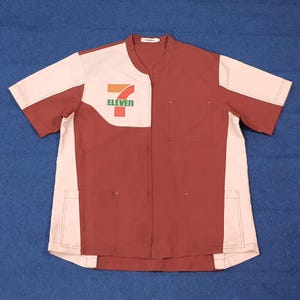 Puede incluir: Una camisa de uniforme de manga corta con el logotipo de "7-Eleven". La camisa es principalmente granate con detalles en crema en las mangas y la parte superior del pecho. Tiene cuello mandarín, cremallera completa y dos bolsillos delanteros.