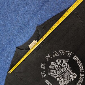 K&ouml;nnte beinhalten: Schwarzes Sweatshirt mit wei&szlig;em "U.S. Navy"-Aufdruck und dem Text "Sports Program". Das Sweatshirt hat einen Rundhalsausschnitt und ein Etikett mit der Aufschrift "AVIREX". Ein gelbes Ma&szlig;band liegt auf dem Sweatshirt.