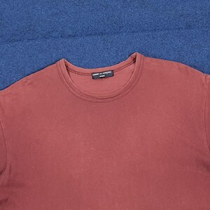 May include: A maroon crew neck t-shirt with the text "COMME des GAR&Ccedil;ONS" printed on the neckline. The shirt is made of a soft material and is displayed on a blue surface.
