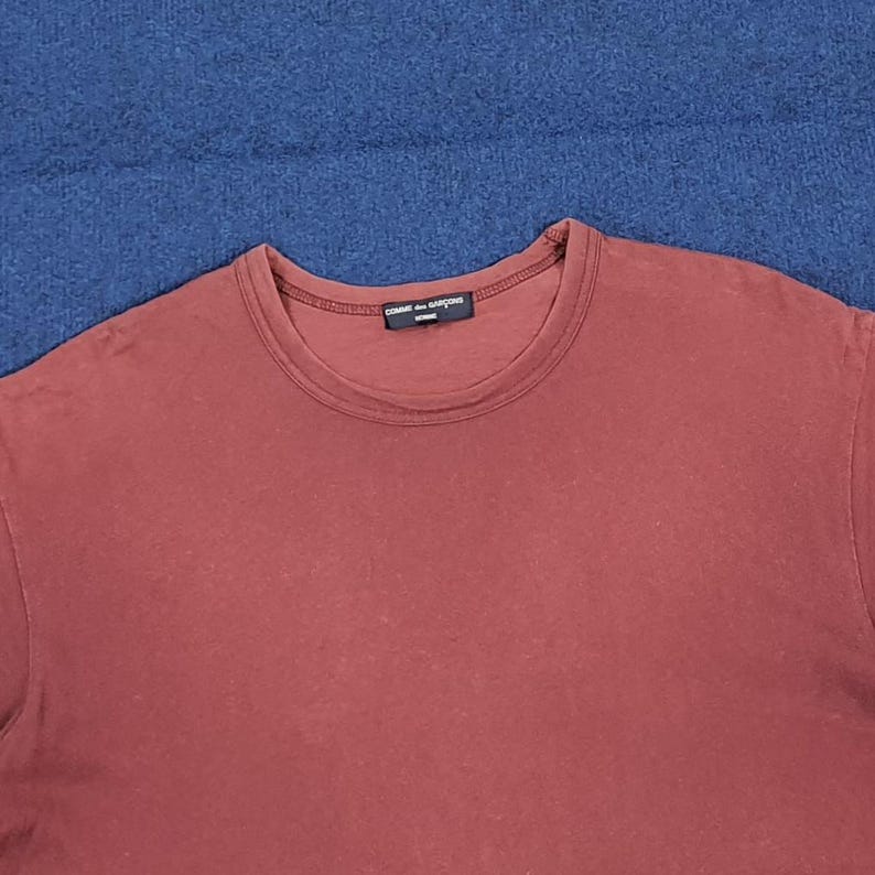 May include: A maroon crew neck t-shirt with the text "COMME des GAR&Ccedil;ONS" printed on the neckline. The shirt is made of a soft material and is displayed on a blue surface.