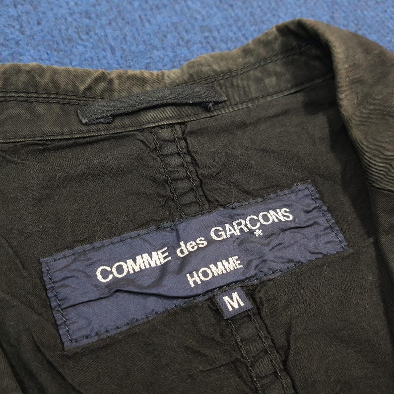 May include: Close-up of a black jacket's interior, featuring a navy blue label with the text "COMME des GARCONS HOMME" in white. The jacket has a size "M" label. The fabric appears worn, with visible stitching and a collar.